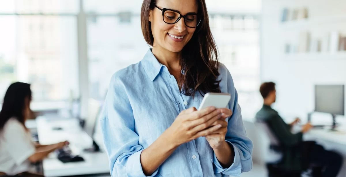 A imagem mostra uma mulher em um escritório mexendo em um smartphone. Ela possivelmente está gerenciando o perfil da empresa e aplicando algumas estratégias para conseguir seguidores no Instagram.