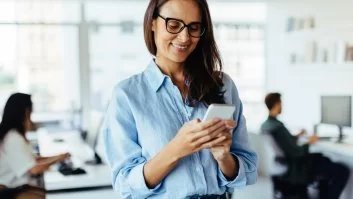 A imagem mostra uma mulher em um escritório mexendo em um smartphone. Ela possivelmente está gerenciando o perfil da empresa e aplicando algumas estratégias para conseguir seguidores no Instagram.