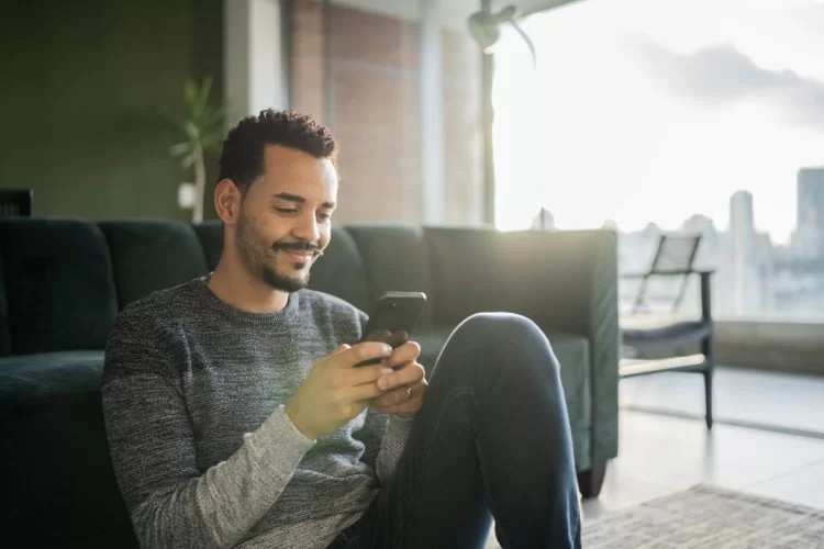 A imagem mostra um homem mexendo em seu smartphone na sala de casa. Ele está sentado no chão e sorri, possivelmente por ter sido impactado por alguma estratégia para conseguir seguidores no Instagram. 