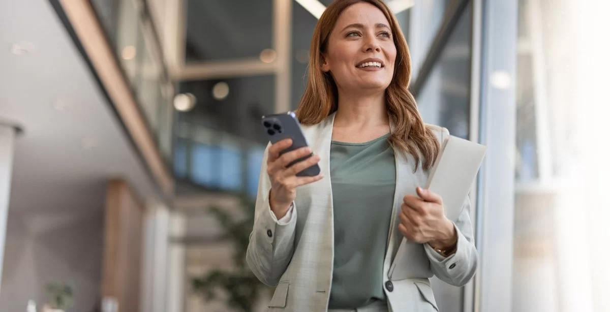 A imagem mostra uma mulher com roupa social no que parece ser o saguão de um escritório. Ela segura o celular em uma mão e uma pasta na outra, olhando para o horizonte enquanto pensa em formas de como monetizar no Instagram.