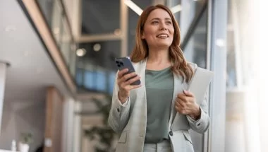 A imagem mostra uma mulher com roupa social no que parece ser o saguão de um escritório. Ela segura o celular em uma mão e uma pasta na outra, olhando para o horizonte enquanto pensa em formas de como monetizar no Instagram.