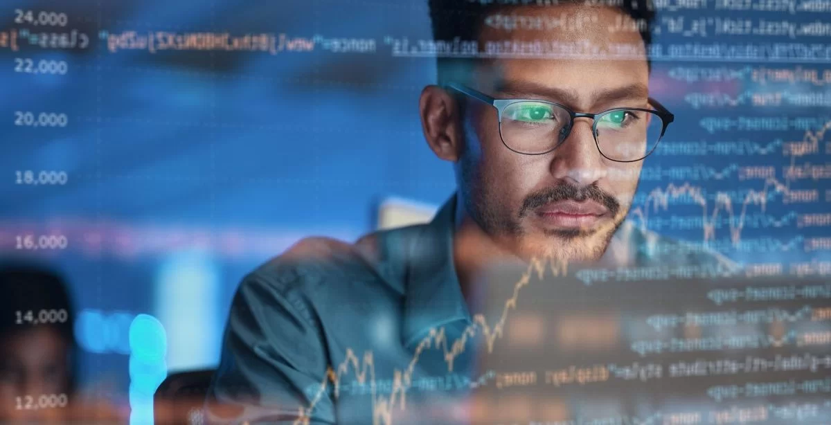 A imagem mostra um homem diante de uma tela com diversas linhas de códigos de programação. Pela quantidade de informações presentes, ele provavelmente está realizando um processo de debug.