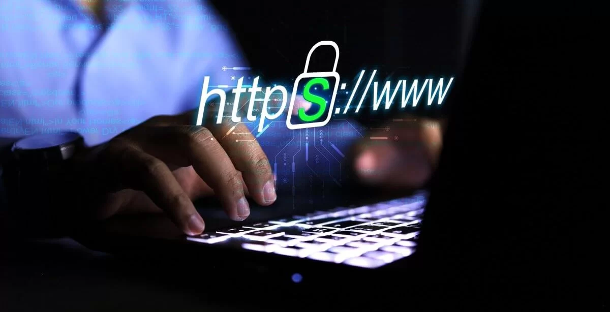 A imagem mostra um homem, possivelmente em um escritório no período noturno, digitando no teclado de um notebook. O rosto dele não aparece. Ele está navegando na internet e acessando um site seguro, pois ele conta com o protocolo HTTPS em sua composição.