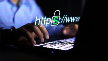 A imagem mostra um homem, possivelmente em um escritório no período noturno, digitando no teclado de um notebook. O rosto dele não aparece. Ele está navegando na internet e acessando um site seguro, pois ele conta com o protocolo HTTPS em sua composição.