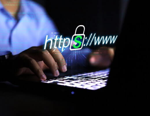 A imagem mostra um homem, possivelmente em um escritório no período noturno, digitando no teclado de um notebook. O rosto dele não aparece. Ele está navegando na internet e acessando um site seguro, pois ele conta com o protocolo HTTPS em sua composição.