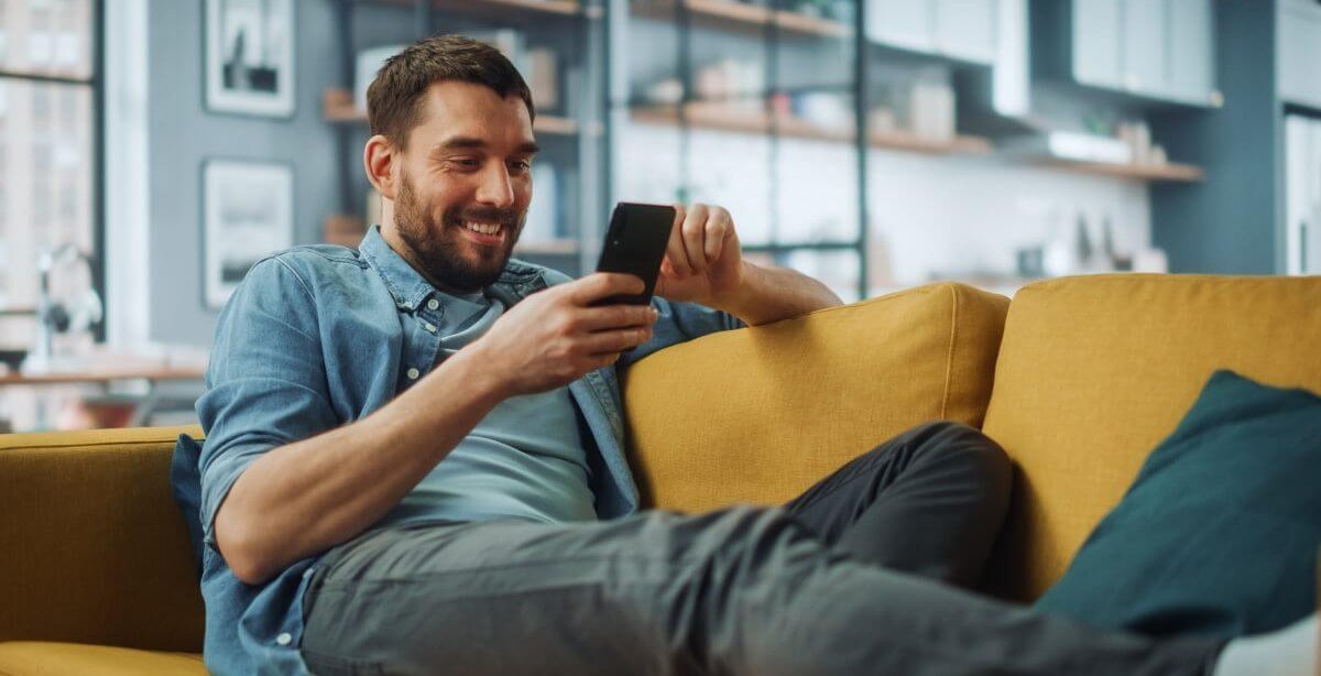 A imagem mostra um homem sorrindo enquanto toca a tela de seu smartphone. Ele possivelmente está em uma rede social e foi impactado pela publicação de uma empresa, que sabe quais são os melhores horários para postar no TikTok.