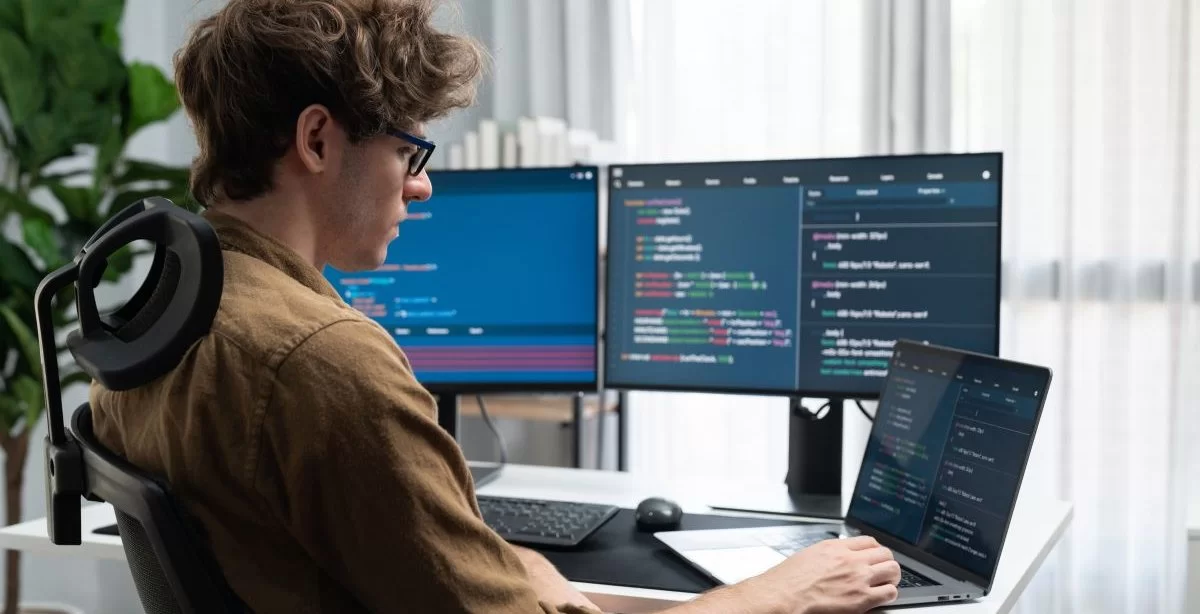A imagem mostra um rapaz branco de óculos trabalhando em um computador. Nas telas, há diversas linhas de código de programação, indicando que ele conseguiu um emprego nessa área após criar e divulgar um portfólio front-end.