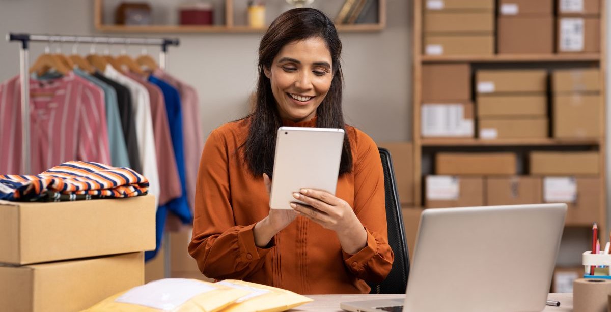 A imagem mostra uma mulher trabalhando em uma loja de roupas que aparentemente é virtual. Ela está segurando um tablet. Há uma mesa com pacotes e um notebook à frente dela, além de uma estante com caixas e uma arara com roupas penduradas.
