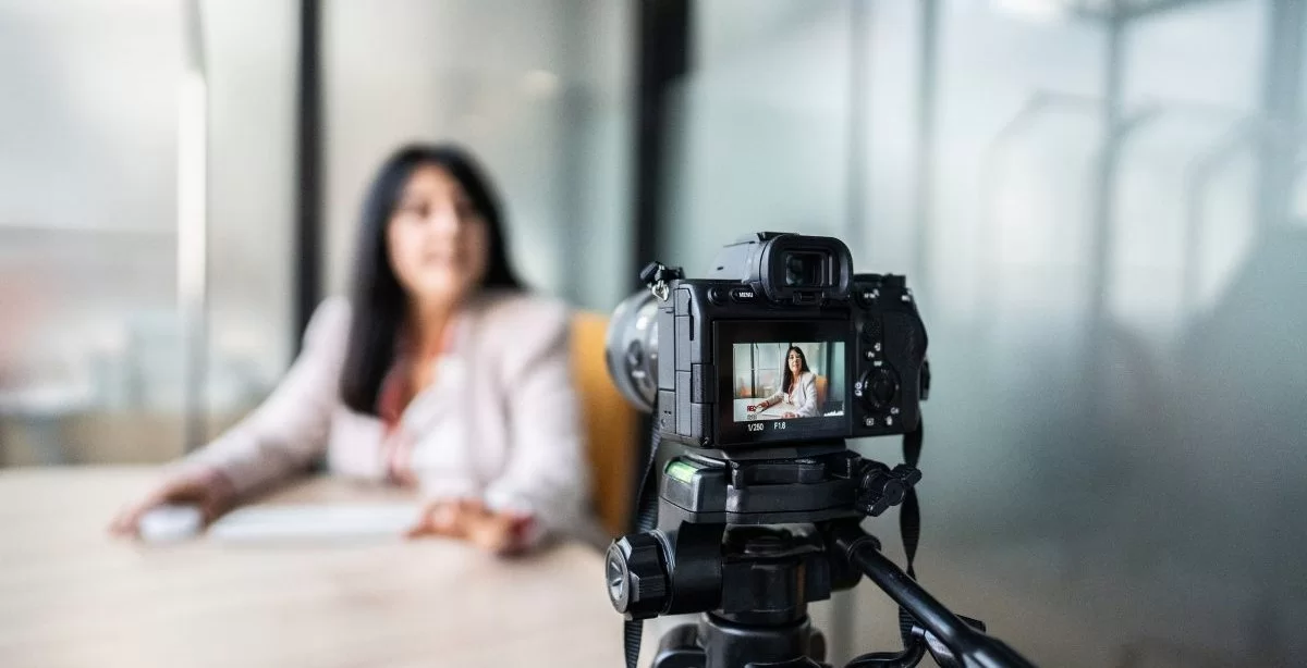 A imagem mostra uma mulher gravando um vídeo com uma câmera. Ela aparece desfocada, já que a foto está mais concentrada na câmera. Ela parece estar gravando um VSL.