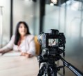 A imagem mostra uma mulher gravando um vídeo com uma câmera. Ela aparece desfocada, já que a foto está mais concentrada na câmera. Ela parece estar gravando um VSL.