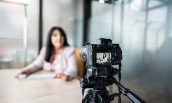 A imagem mostra uma mulher gravando um vídeo com uma câmera. Ela aparece desfocada, já que a foto está mais concentrada na câmera. Ela parece estar gravando um VSL.