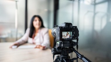 A imagem mostra uma mulher gravando um vídeo com uma câmera. Ela aparece desfocada, já que a foto está mais concentrada na câmera. Ela parece estar gravando um VSL.