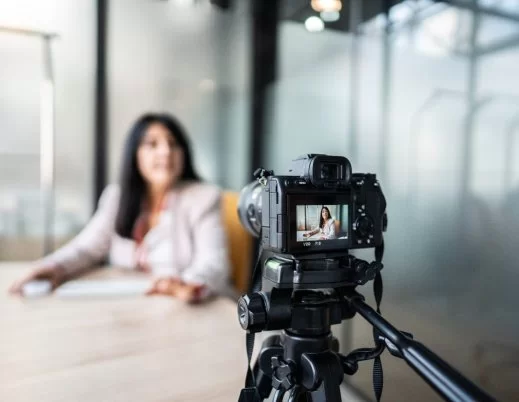 A imagem mostra uma mulher gravando um vídeo com uma câmera. Ela aparece desfocada, já que a foto está mais concentrada na câmera. Ela parece estar gravando um VSL.