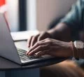 As mãos de uma pessoa afro-americana estão digitando no teclado de um notebook, possivelmente o endereço de um site que possa ser .com ou .com.br.
