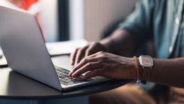 As mãos de uma pessoa afro-americana estão digitando no teclado de um notebook, possivelmente o endereço de um site que possa ser .com ou .com.br.