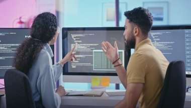 Dois jovens programadores, sendo um homem branco, de costas, e uma mulher afro-americana, de lado; ambos vestindo roupas casuais, estão de frente para monitores observando linhas de código e usando Flask Python.