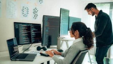 A imagem mostra um homem e uma mulher conversando na frente de um computador com várias telas. Há alguns códigos nelas, e eles possivelmente estão debatendo sobre formas de aplicar o Machine Learning na empresa onde trabalham.