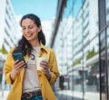 A imagem mostra uma mulher andando pela rua, enquanto segura um smartphone com uma mão e um copo de café com a outra. Ela provavelmente foi impactada por uma publicação em uma rede social, possivelmente feita por uma empresa que estudou qual é o melhor horário para postar no Instagram.