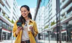 A imagem mostra uma mulher andando pela rua, enquanto segura um smartphone com uma mão e um copo de café com a outra. Ela provavelmente foi impactada por uma publicação em uma rede social, possivelmente feita por uma empresa que estudou qual é o melhor horário para postar no Instagram.