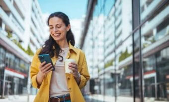 A imagem mostra uma mulher andando pela rua, enquanto segura um smartphone com uma mão e um copo de café com a outra. Ela provavelmente foi impactada por uma publicação em uma rede social, possivelmente feita por uma empresa que estudou qual é o melhor horário para postar no Instagram.