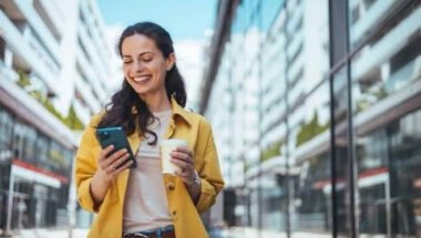 A imagem mostra uma mulher andando pela rua, enquanto segura um smartphone com uma mão e um copo de café com a outra. Ela provavelmente foi impactada por uma publicação em uma rede social, possivelmente feita por uma empresa que estudou qual é o melhor horário para postar no Instagram.