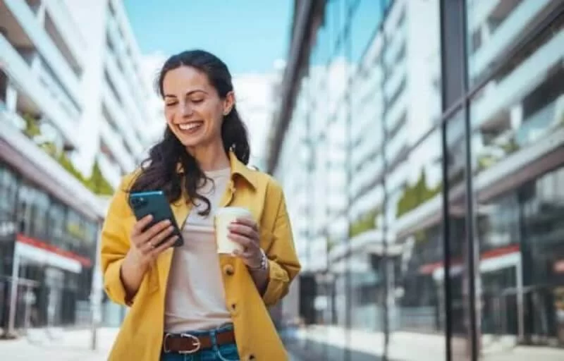 A imagem mostra uma mulher andando pela rua, enquanto segura um smartphone com uma mão e um copo de café com a outra. Ela provavelmente foi impactada por uma publicação em uma rede social, possivelmente feita por uma empresa que estudou qual é o melhor horário para postar no Instagram.