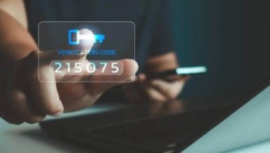 A imagem mostra uma pessoa segurando um smartphone com uma mão e tocando uma tela de código de verificação com a outra. Esse é um procedimento padrão que representa as ações da verificação em duas etapas.
