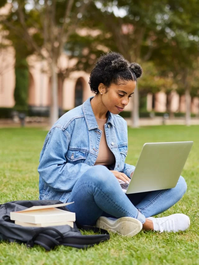 Uma mulher jovem negra está sentada em um parque com um notebook no colo, vestindo roupas casuais. A imagem pode representar o conceito de TI verde.