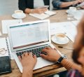 Um homem está acessando uma página na internet em seu notebook, posicionado em uma mesa com uma xícara de café, óculos, smartphone e papeis ao redor. A página virtual acessada por ele possui em sua URL um favicon.