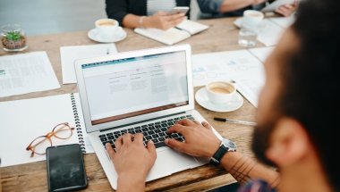 Um homem está acessando uma página na internet em seu notebook, posicionado em uma mesa com uma xícara de café, óculos, smartphone e papeis ao redor. A página virtual acessada por ele possui em sua URL um favicon.
