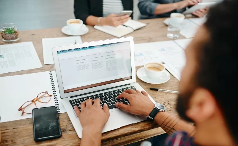 Um homem está acessando uma página na internet em seu notebook, posicionado em uma mesa com uma xícara de café, óculos, smartphone e papeis ao redor. A página virtual acessada por ele possui em sua URL um favicon.