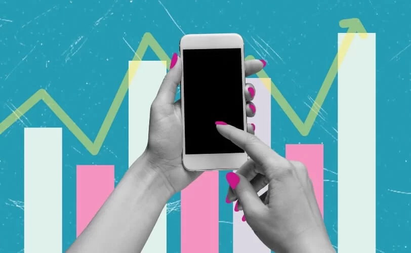 Um par de mãos femininas desliza um dos dedos indicadores sobre a tela do celular. Ao fundo, é possível visualizar barras brancas e rosas, que compõe um gráfico disposto sob as mãos, representando a ideia em torno das principais tendências de marketing para 2025.