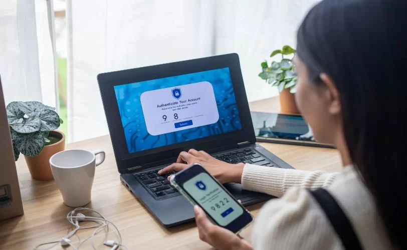 Uma pessoa, cujas mãos aparecem na imagem, está utilizando um notebook e um celular para realizar a verificação em duas etapas, uma medida de segurança para proteger seus dados pessoais de acessos não autorizados. 