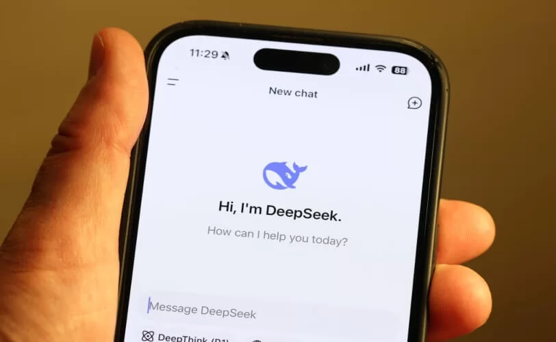 Imagem mostra um smartphone exibindo o logotipo do "DeepSeek", sendo segurado por uma mão de uma pessoa branca. O design sugere uma identidade visual moderna e tecnológica, relacionada a uma ferramenta de inteligência artificial.