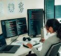 Uma jovem mulher de perfil, vestindo suéter cinza e camisa branca, está em frente a dois monitores de computador e um notebook. Ela digita linhas de código e avalia um a um, usando uma linguagem de programação.