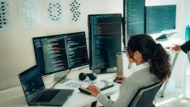 Uma jovem mulher de perfil, vestindo suéter cinza e camisa branca, está em frente a dois monitores de computador e um notebook. Ela digita linhas de código e avalia um a um, usando uma linguagem de programação.