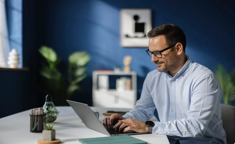 Um homem branco, usando camisa azul-claro, óculos de grau de aro preto e relógio, digita em um notebook cinza. O equipamento está sobre uma mesa branca e, ao seu lado, está um tablet de capa verde. Em sua frente, há um porta-lápis com materiais de escritório e objetos de decoração