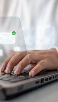 Uma pessoa branca, usando camisa azul claro e gravata cinza, digita em um notebook. Sobre a fotografia, surgem ícones representando e-mails que se conectam entre si.