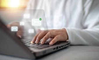 Uma pessoa branca, usando camisa azul claro e gravata cinza, digita em um notebook. Sobre a fotografia, surgem ícones representando e-mails que se conectam entre si.