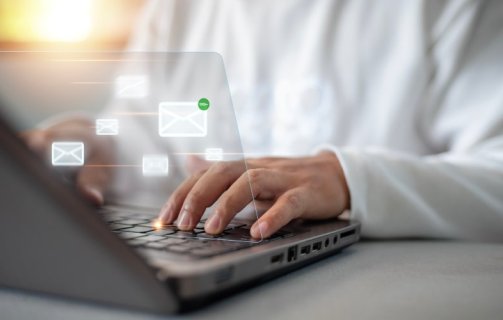 Uma pessoa branca, usando camisa azul claro e gravata cinza, digita em um notebook. Sobre a fotografia, surgem ícones representando e-mails que se conectam entre si.