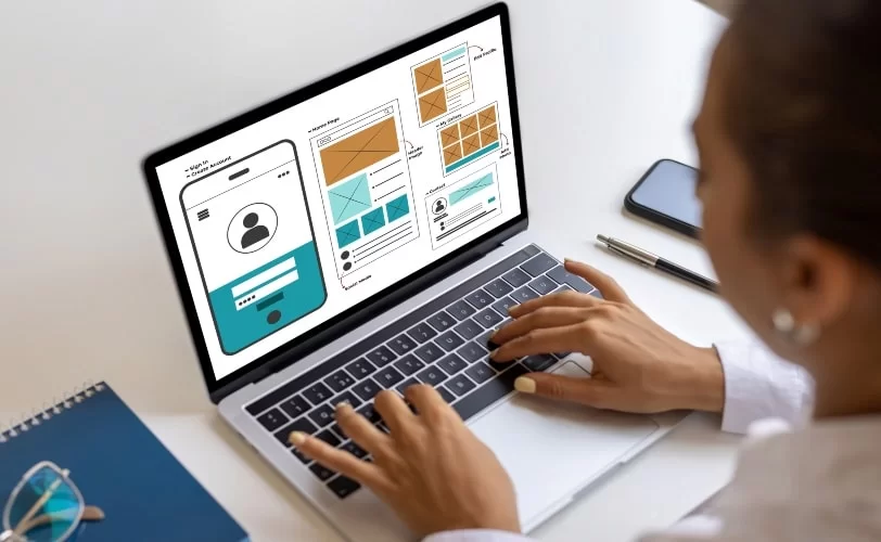 A imagem mostra uma profissional trabalhando no design de interface de um site ou aplicativo em um laptop. A tela do computador exibe wireframes e layouts estruturados para páginas web e mobile, com seções organizadas para perfis de usuário, galerias de imagens e redes sociais. O ambiente de trabalho inclui um caderno, uma caneta e um smartphone, sugerindo um processo criativo e produtivo de design UX/UI, tudo para evitar criar sites não responsivos. 