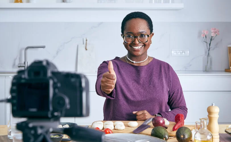 A imagem mostra uma mulher negra gravando um vídeo com uma câmera fotográfica. Ela aparentemente está na cozinha produzindo material sobre culinária, pois há ingredientes na mesa. Após essa produção, ela possivelmente usará a Inteligência Artificial para auxiliar na pós-produção e em aspectos mais criativos.