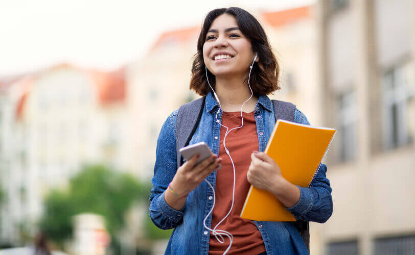 Uma mulher jovem está na rua usando seu smartphone enquanto veste camisa jeans, blusa marrom e fones de ouvido. 
