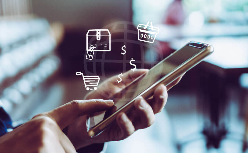 Pessoa segurando smartphone com ícones de e-commerce flutuando na tela, representando compras online, pagamento digital e carrinho de compras.