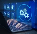 A imagem mostra uma cena conceitual de automação e tecnologia digital. Um profissional está digitando em um notebook, enquanto sobre a tela aparecem ícones digitais em azul, representando diferentes processos: engrenagens, checklist, segurança, inteligência artificial e validações. O destaque principal é o ícone de engrenagens em movimento, cercado por setas circulares, simbolizando o fluxo contínuo de automação de tarefas.