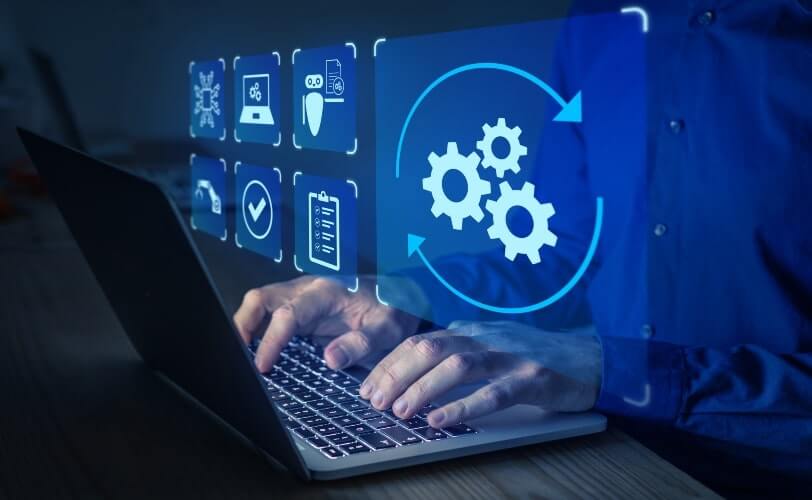 A imagem mostra uma cena conceitual de automação e tecnologia digital. Um profissional está digitando em um notebook, enquanto sobre a tela aparecem ícones digitais em azul, representando diferentes processos: engrenagens, checklist, segurança, inteligência artificial e validações. O destaque principal é o ícone de engrenagens em movimento, cercado por setas circulares, simbolizando o fluxo contínuo de automação de tarefas.