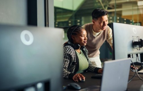 Dois desenvolvedores, homem e mulher, analisando código em um monitor em ambiente de trabalho escuro