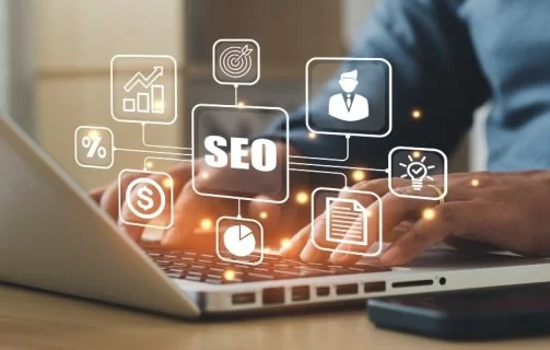 Pessoa digitando em um notebook com ícones digitais flutuantes relacionados a SEO, como gráfico de crescimento, alvo, perfil de usuário, lâmpada de ideia e cifrão, representando estratégias de otimização para mecanismos de busca e performance online.