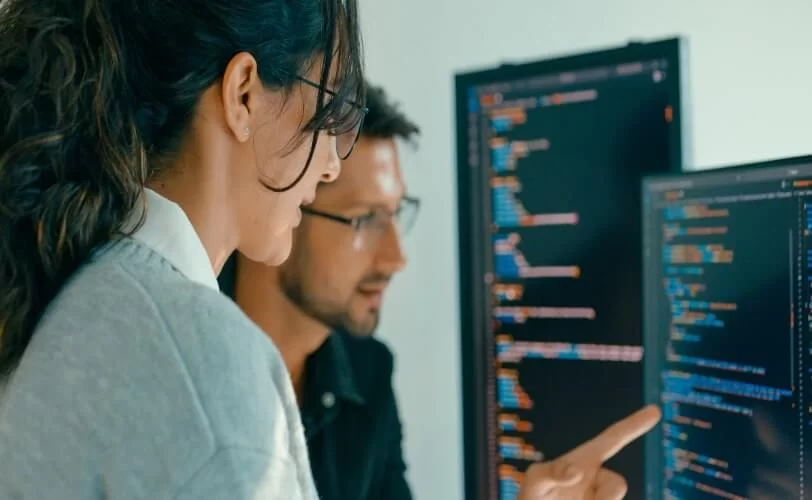 Dois profissionais de tecnologia, sendo um homem e uma mulher, trabalham juntos em frente a monitores com códigos coloridos. Em um ambiente escuro e moderno, a mulher aponta para a tela vertical enquanto o homem observa atentamente, indicando uma colaboração ativa na análise ou desenvolvimento de software.