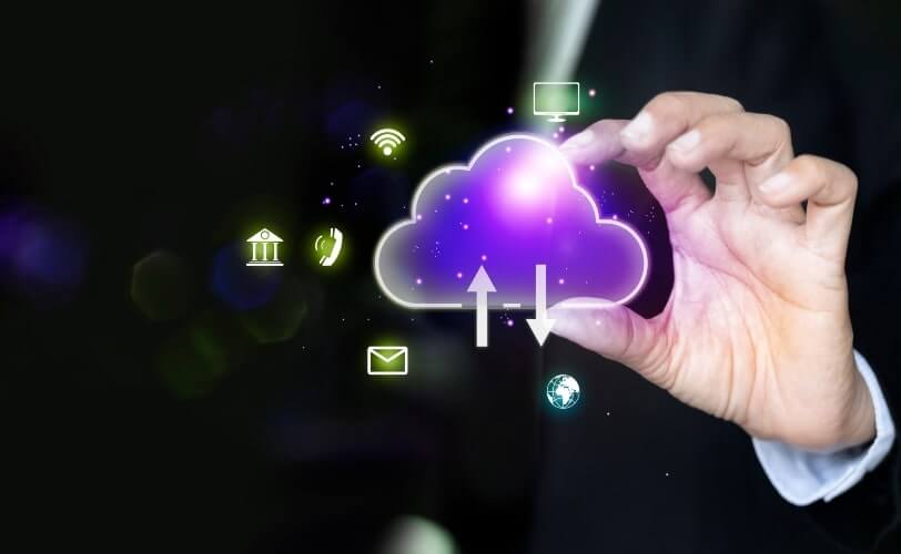 A imagem apresenta um ícone de nuvem digital com setas de upload e download, simbolizando o armazenamento e compartilhamento de informações em sistemas de computação em nuvem. As linhas luminosas e o fundo azul com partículas tecnológicas reforçam a ideia de fluxo de dados, conectividade e infraestrutura digital. Essa representação ilustra como a nuvem (cloud computing) permite o acesso remoto, a integração entre dispositivos e a agilidade nos processos corporativos, sendo um pilar essencial da transformação digital e da inovação tecnológica.